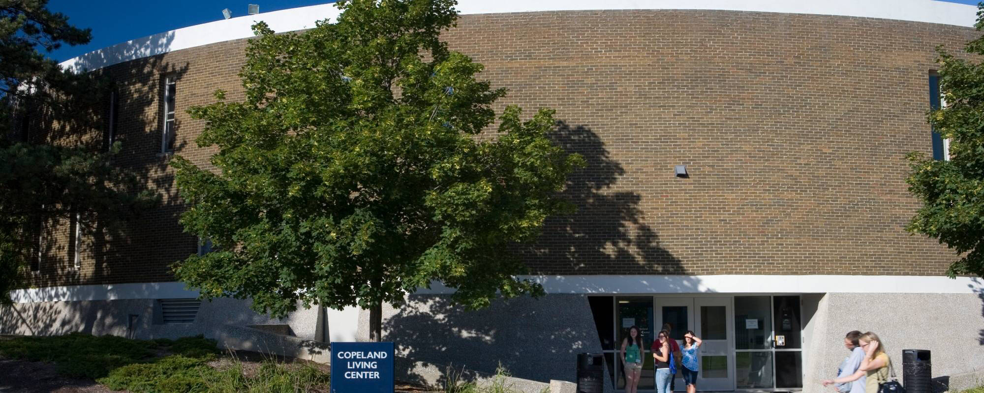 A brick building labeled "Copeland Living Center" has a tree casting shadows on its facade. Three people stand outside the entrance, engaged in conversation.